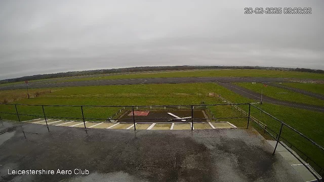 A view from a webcam showing an airport environment at Leicestershire Aero Club. The scene is overcast with a gray sky and a hint of fog. In the foreground, there is a railing, and below it, a small area is visible with a white plane resting on the ground. The background features a large green grassy field and a paved runway. Several signs are positioned along the edge of the runway, and some trees can be seen in the distance. The setting appears quiet and tranquil, suggesting a calm day at the airport.