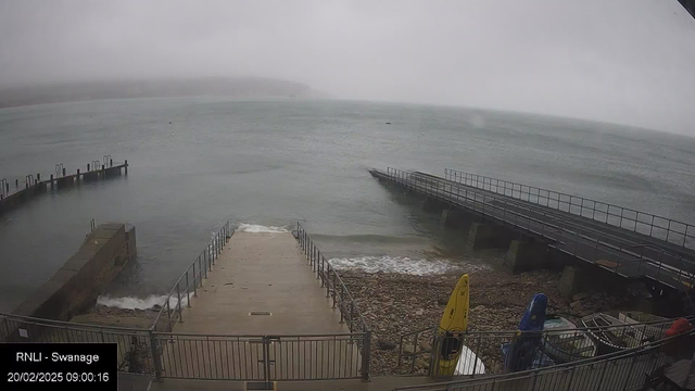 A foggy seaside scene showing a concrete ramp leading down to a rocky shoreline, with waves lapping at the edge. Two docks are visible: one to the left with pilings and one to the right extending out over the water. Several brightly colored kayaks, in yellow and blue, are positioned on the right side near the shoreline. The sky is overcast, creating a grayish atmosphere over the water.