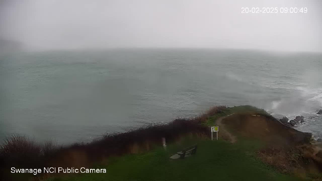 The image shows a coastal scene with choppy waters under overcast skies. There is heavy mist or rain reducing visibility. In the foreground, there is a grassy area with a bench and a warning sign. The shoreline curves gently, leading into rolling waves that crash against the rocks. The overall atmosphere appears stormy and chilly.