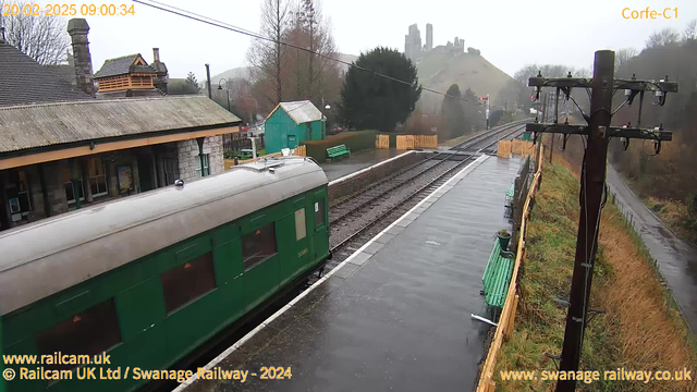 A foggy scene at a train station featuring a green train car partially in view on the left side. The platform is lined with benches, and there are trees and bushes in the background. On the right, a power pole with wires stands beside the train tracks, which appear wet. In the distance, a hillside rises with castle ruins. The atmosphere is overcast and damp, suggesting rainy weather.