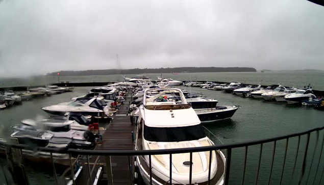 A marina with several boats docked in water under a cloudy sky. The scene is slightly rainy, with a grayish tone dominating the view. Various types of boats are moored, some with white hulls, while others are darker. A wooden dock runs along the edge of the marina, and there are faint outlines of land in the background. Overall, the atmosphere is misty and quiet, typical of a cool, overcast day by the water.