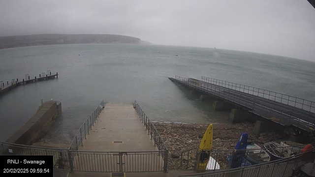 A cloudy scene at Swanage shows a wide, gray sea with gentle waves. In the foreground, a concrete ramp leads to the water, bordered by railings. To the left, there is a small wooden pier extending into the sea, while a larger, wider pier is visible on the right. Several colorful kayaks, including yellow and blue, are stored on the shore near some rocks, indicating a waterfront area. The distant shoreline is partially obscured by mist, with cliffs rising in the background.