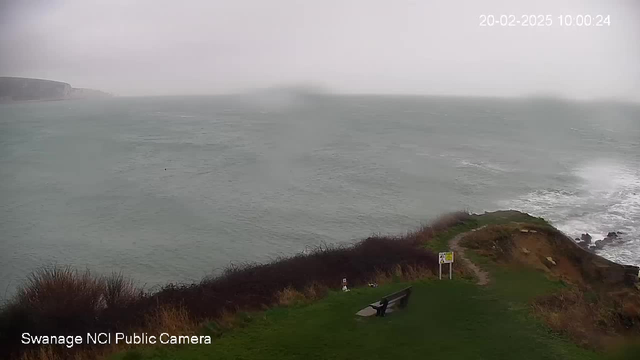 A coastal scene during overcast weather, featuring rough sea waves crashing against a rocky shore. In the foreground, there is a grassy area with a bench positioned to the right. A path leads from the grassy area towards the edge of the cliff. To the right, a sign is visible amidst the vegetation. The horizon is blurred due to misty conditions. Overall, the atmosphere appears gloomy and windy.