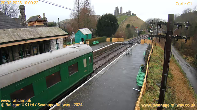 A green train car is seen on the left side of the image, parked at a train station. The station platform is wet, likely from recent rain. In the background, there is a small green building. Beyond that, the landscape rises to a hill where the ruins of a castle are visible. Wooden benches line the platform, and there are overhead power lines above the scene. The sky appears overcast, giving a gray ambiance to the setting.