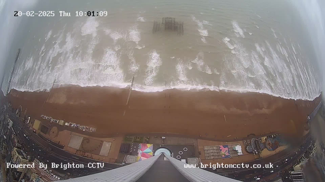 Aerial view of a beach with sandy shore and waves crashing against it. In the distance, a pier structure is partially submerged in water. The beach appears to have colorful patterns and objects along the shoreline. Above the scene, there is a cloudy sky, and the image includes a timestamp and a watermark indicating it is powered by Brighton CCTV. The overall atmosphere is tranquil, with no visible people on the beach.