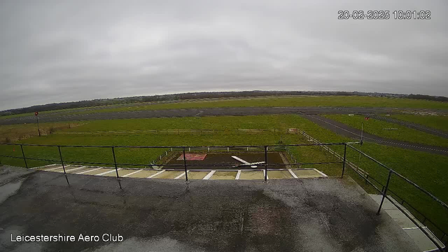 A view from a webcam at Leicester Aero Club. The foreground shows a wet concrete area with a railing. In the background, there is a wide green field and an airstrip lined with dark asphalt. The sky is overcast and gray, indicating cloudy weather. Some red and white markers are visible along the runway, and a partial view of the landscape can be seen in the distance.