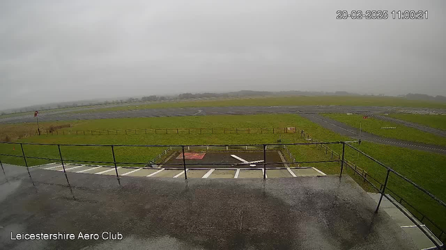 A wide view of a gray, overcast landscape taken from a raised platform. In the foreground, there is a railing and a flat surface, possibly a terrace. Below, there's a grassy area with a red and white sign, alongside a small tarmac section that appears wet. The horizon features a patch of grass and a distant tree line, all under a cloudy sky, indicating rainy weather. The scene lacks any visible aircraft.