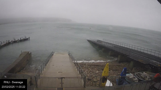 A view of a misty waterfront scene at Swanage, with a calm sea and a grey sky. In the foreground, there are two jetties extending into the water; one is wider and made of concrete, while the other is narrow with railings. The water is lapping at the base of the jetties, and along the shore, there are smooth pebbles. A few colorful boats, including a yellow kayak and a blue boat, are positioned on the right side near the shore. The overall atmosphere is quiet and serene, with a faint silhouette of distant land in the background.