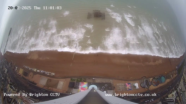 Aerial view of a beach with waves crashing on the shore. The sandy beach is visible, along with a section of a boardwalk. A pier structure is located in the water, partially submerged. Several colorful attractions and buildings line the beach area, including a large circular feature. The scene is set on a cloudy day, with gray skies and ocean water appearing turbulent. A digital timestamp is displayed at the top left corner.