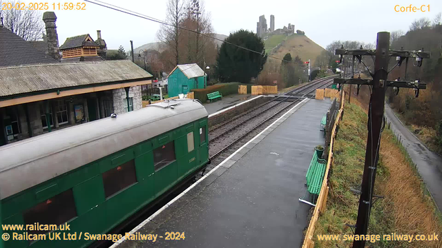 A green train car is positioned to the left, partially off-screen. The setting is a railway station with wet, reflective pavement. In the background, there is a hill with castle ruins. Station buildings with a rustic design can be seen, along with a small, green shed and benches along the platform. There are railway tracks that lead into the distance. The sky appears overcast.