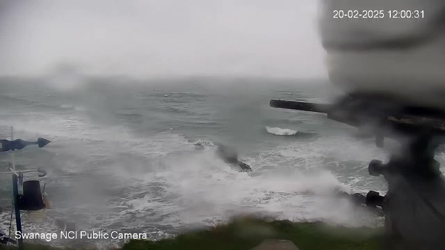 A seaside view during a stormy day showing rough waves crashing against rocks. The sky is cloudy and gray, indicating inclement weather. There is a blurred structure in the foreground, likely a camera or observation equipment. The scene conveys a sense of turbulence and dynamic movement in the ocean.