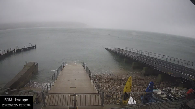 A foggy seaside scene showing a wide body of water with rocky shoreline in the foreground. A concrete ramp leads down to the water, flanked by a railing. On the right, a wooden pier extends into the water, partially obscured by fog. Several boats are visible near the shore, including yellow and blue kayaks. The background shows a hazy landscape, blending into the water. The timestamp in the corner indicates the date and time of the image.