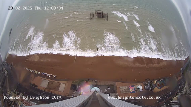 A webcam view looking straight down from a height above the ocean. Below, the brown sandy beach is visible, with waves crashing onto the shore. There is an old, partially submerged wooden pier in the water. To the sides, there are structures and people on the beach, as well as a colorful playground area seen on the sand. The sky appears cloudy, and the timestamp indicates the image is taken on February 20, 2025, at noon.