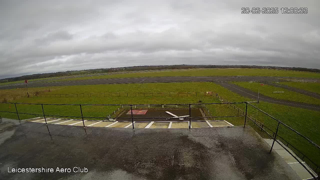 A webcam view from the Leicestershire Aero Club, showing a wide expanse of green grass and a runway. The sky is overcast with gray clouds, and there are a few scattered structures on the field. A low railing is visible in the foreground, and a red sign can be seen in the distance near the runway. The ground appears wet, indicating recent rain.