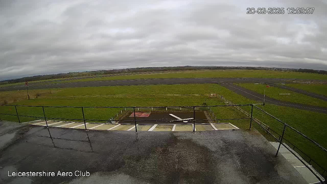 A view from a webcam at Leicestershire Aero Club, showing a wide expanse of a grassy airfield under a cloudy sky. In the foreground, there is a railing, likely from a viewing platform, with some wet surfaces visible. The airstrip stretches across the middle of the image, with an empty runway and a small red flag in the background. The scene conveys a muted, overcast atmosphere with no visible aircraft or people.