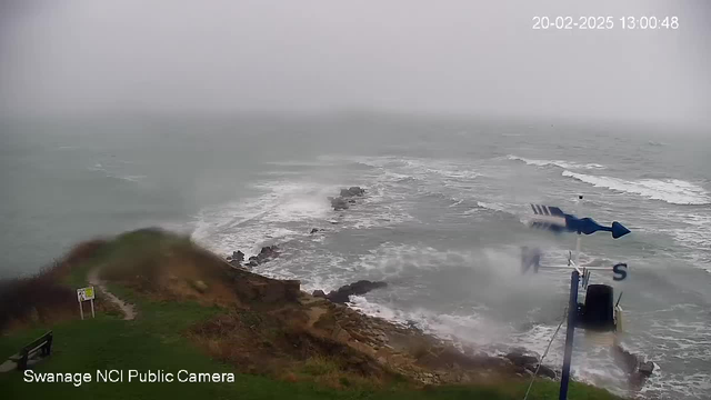 A coastal scene with rough seas and waves crashing against rocky formations. The sky is overcast and gray, indicating stormy weather. In the foreground, a grassy area slopes down to the water, with a bench visible on the left side. A sign is also present near the edge. A weather vane stands on the right, showing the direction of the wind. The date and time are visible in the top right corner, indicating a date in February 2025.