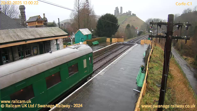 A green train car is partially visible on the left side of the image, sitting on a wet railway platform. The railway tracks extend to the right, where there is a wooden fence and some greenery in the background. In the distance, a hill is visible with the ruins of a castle. The sky is overcast with gray clouds, and there are benches along the platform. On the right side, a wooden utility pole with wires is present. The scene appears to be a rural train station.