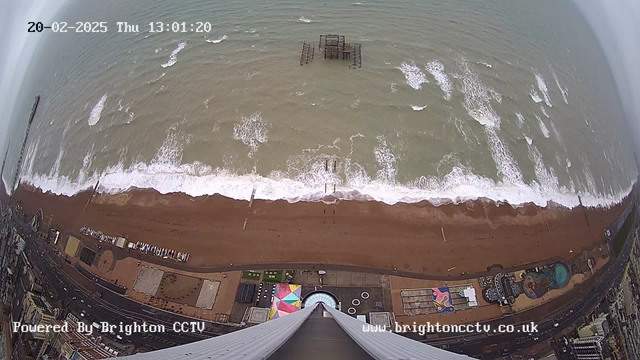 Aerial view of a beach with crashing waves and a sandy shoreline. In the center, there is a partially submerged pier structure. The bottom part of the image shows elements of a beachfront promenade with colorful patterns on the ground and various facilities. The sky appears overcast. The timestamp reads "20-02-2025 Thu 13:01:20." The image is watermarked with "Powered by Brighton CCTV."