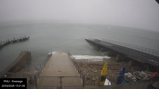 The image shows a foggy coastal scene with a view of the water and a sloped ramp leading down to the shore. In the foreground, there is a concrete path with a railing on either side. To the right, several colorful kayaks are parked, including yellow and blue. A wooden pier extends over the water, with some structures visible on the far side. The water appears calm with gentle waves lapping at the shore, and the visibility is limited due to the fog in the background.