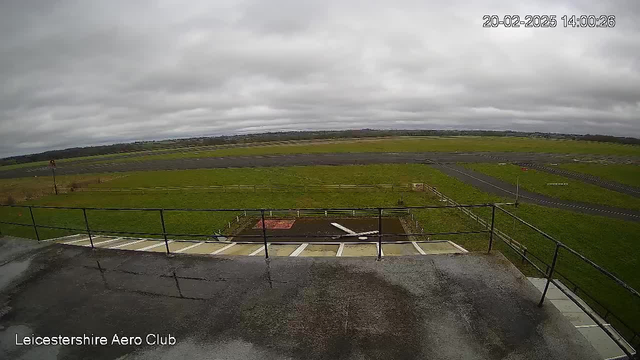 A cloudy sky fills the top of the image. Below, a wide open field of green grass stretches across the scene, with a gray runway in the middle. There are several wooden fences bordering the runway. In the foreground, a railing can be seen, suggesting a viewpoint or balcony area. In the distance, a small tree line is visible against the horizon. The date and time are displayed in the corner of the image. The ground appears wet and reflective.