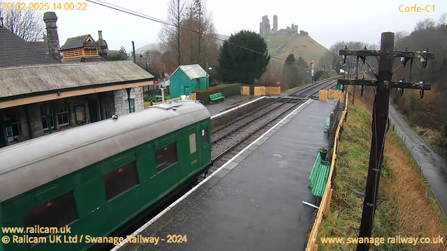 A green train carriage is positioned on a wet train platform. In the background, a station building with a stone facade and a sloped roof is visible. To the right, there are several green benches along the platform. Beyond the station, a hilly area with rocky ruins is partly obscured by mist. A wooden fence separates the platform from the grassy hillside, and a power pole stands near the edge of the image. The sky is overcast, indicating a cloudy day.