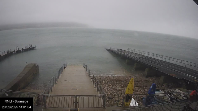 A foggy seascape at Swanage with gentle waves lapping against a rocky shoreline. In the foreground, a wide concrete ramp leads down to the water, flanked by a railing. To the right, a small dock extends into the water, lined with metal posts. Kayaks in yellow and blue are visible on the shore, along with some scattered rocks. The scene is dim, shrouded in mist, with a muted color palette.