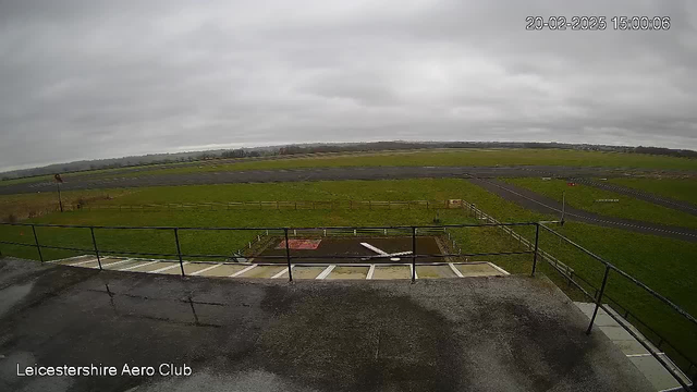 A cloudy day at the Leicestershire Aero Club. The image shows a grassy field with a tarmac runway in the foreground, bordered by a wooden fence. A slight overhang with a railing is visible in the bottom part of the image. The runway is empty, and the landscape is mostly flat with distant trees lining the horizon. The sky is overcast and gray, indicating a dreary atmosphere.