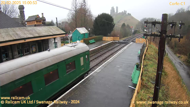 A green train carriage is visible on the left side of the image, with a cloudy, grey sky above. To the right, a railway track runs parallel alongside a platform, with wooden benches in a green color positioned on the platform. In the background, there are trees and a hill featuring ruins, partially obscured by mist. The setting appears to be a rural railway station, and the overall atmosphere is overcast and somewhat desolate.