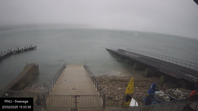 A misty waterfront scene shows calm water with limited visibility. In the foreground, a wide concrete slipway leads down to the water, bordered by metal railings. To the right, a wooden pier extends into the water, partially obscured by fog. On the shore, several colorful kayaks and small boats are stacked near the water's edge, with smooth stones visible along the shoreline. The sky is overcast, contributing to the hazy atmosphere. A timestamp indicates the date and time of the image.