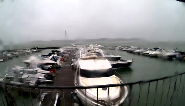 A view of a marina on a cloudy and rainy day. Several boats are docked in the water, with some covered by tarps and others visible in various shapes and sizes. The atmosphere is overcast, with water droplets on the camera lens causing some blurriness in the image. In the background, a faint outline of land can be seen, and there is a bridge in the distance, partially obscured by the weather.