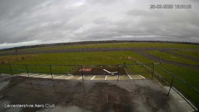 A cloudy sky stretches over a grassy airfield. In the foreground, there is a railing along a low platform, with a wet surface indicative of recent rain. Below, the airfield has grassy patches and marked runways, with a few scattered objects and signs. The overall atmosphere is calm, with no visible aircraft or people in sight.