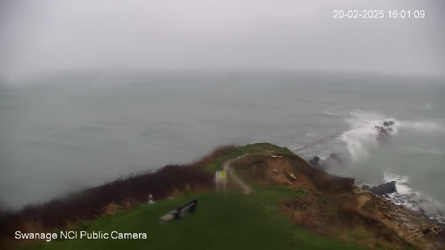 A coastal scene shows a partially obscured view of the sea under overcast skies. Waves crash against a rocky shore in the distance. The foreground includes a grassy area with a path leading towards a white and yellow sign. Nearby, there are two dark benches facing towards the sea. The image is blurred, indicating poor visibility due to weather conditions. The timestamp in the corner shows the date and time.