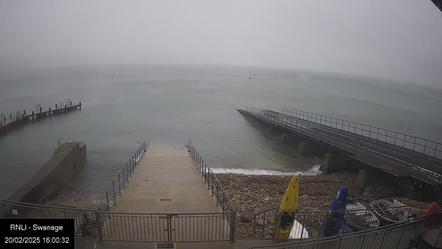 A view of a harbor on a misty day. The water appears calm with faint ripples and is surrounded by a gray sky. To the left, there is a concrete pier with several boat mooring posts visible. A second, wider concrete ramp leads down to the water, and at the bottom of the ramp, there are stones. On the right, a wooden ramp extends into the water. Nearby, there are various colored kayaks, including yellow and blue, resting on the shore. The scene has a serene yet overcast atmosphere.