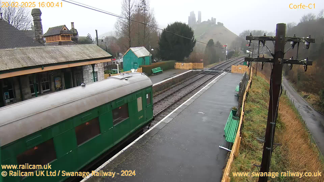 A green train carriage is positioned on the left side of the image, with its roof partially visible. The setting is a train station with a grey, foggy atmosphere. To the right, railway tracks extend into the distance, leading toward a hill where partially ruined stone structures can be seen at the top. The area has benches painted in green, a wooden fence, and various trees and shrubs in the background. The image conveys a quiet, rural station environment.