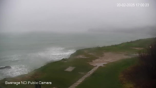 A gray, overcast sky looms over a rough sea with choppy waves crashing along a rocky shoreline. The foreground shows a grassy area with a path leading towards the water. The visibility is low due to fog, and the scene appears quite gloomy, suggesting stormy weather. A timestamp in the corner indicates the date and time as 20th February 2025 at 17:00:23.