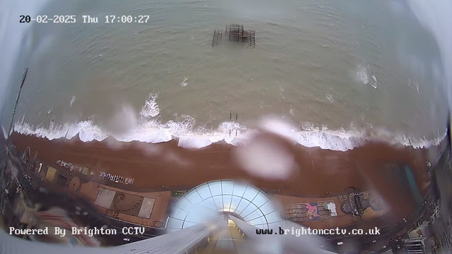 A view from a high vantage point overlooking the coastline. The sandy beach is visible, with white waves crashing onto the shore. In the water, remnants of a wooden pier can be seen. The sky appears overcast, likely a dull day. Information is displayed at the top with the date and time. The image is slightly obscured by water droplets, which may indicate rain. The area has some structures and decorations along the beach.