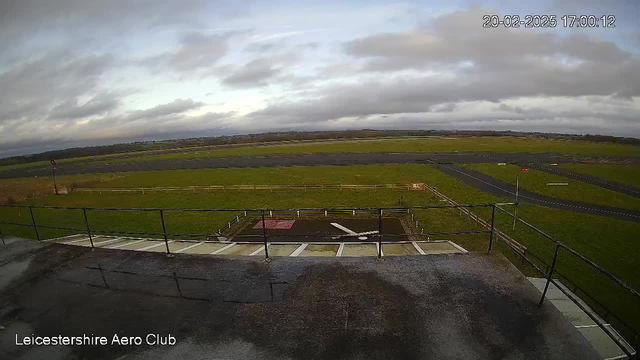 A view from a webcam overlooking a grassy field and runway of the Leicestershire Aero Club. The sky is partly cloudy with gray clouds, and the ground is wet, possibly from recent rain. There are wooden fences in the foreground, and an area of the runway is visible in the background, with markings and a few parking areas. The image is timestamped February 20, 2025, at 17:00:12.