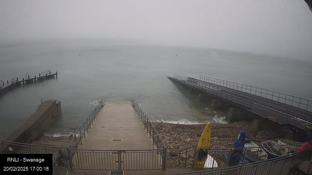 A foggy waterfront scene with a view of the sea and a shoreline. There are two piers extending into the water; one is made of concrete and the other appears to be a metal structure. A wide, concrete ramp leads down to the water, bordered by metal railings. In the foreground, there are several kayaks in different colors, including yellow and blue, resting on the rocky shore. The sky is mostly gray and overcast, contributing to a muted color palette throughout the image.