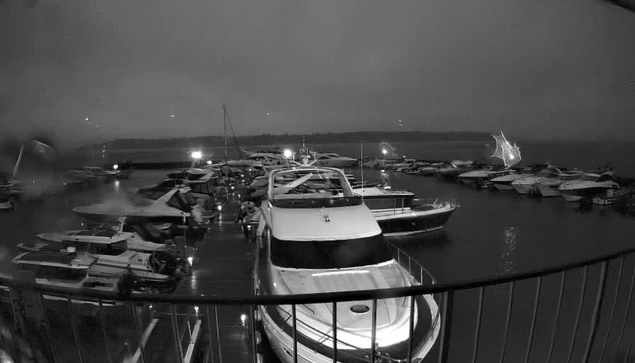 A black and white image of a marina during nighttime. Several boats are docked at the pier, with their silhouettes visible against the dark water. Some have lights shining, creating reflections in the water. The scene has a foggy or damp atmosphere, with limited visibility in the distance where the shoreline fades into darkness.