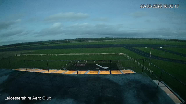 A webcam view of an open field and runway. In the foreground, there is a balcony with a flat surface, partially visible. The ground is predominantly green grass, with a few white markers scattered across. A black asphalt runway extends across the middle of the image, bordered by a white fence. The sky is overcast with gray and blue clouds, indicating a cloudy day.