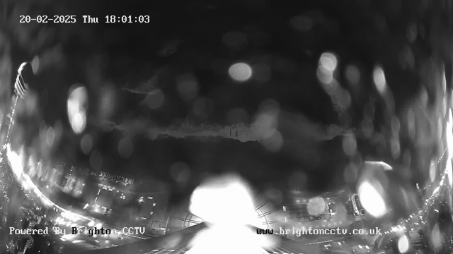 A black and white image from a webcam showing a rainy city street at dusk. Raindrops are visible on the lens, creating a blurry effect. Streetlights and vehicle headlights are faintly illuminated, reflecting off the wet pavement. In the background, dark clouds are present in the sky. The date and time displayed in the corner indicate it is February 20, 2025, at 18:01:03. A watermark at the bottom reads "Powered By Brighton CCTV" and includes a website link.