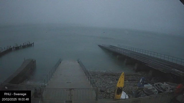 A misty scene at a waterfront with a ramp leading down to the water. On the left, a concrete structure extends into the sea, and several metal posts are visible at the end. The water appears calm and is a gray-blue color, blending with the overcast sky. On the right, yellow and blue boats are secured on the shore, surrounded by a rocky area. The overall atmosphere is foggy and subdued, suggesting a tranquil, yet somber setting.