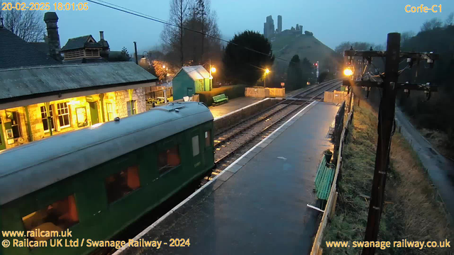 A foggy evening scene at Corfe Castle train station. In the foreground, a green train carriage is partially visible, parked on the tracks. The station building, illuminated with warm lights, shows large windows and a sloping roof, with a green shed nearby. There are wooden benches along the platform and illuminated lampposts. In the background, Corfe Castle's ruins are visible atop a hill, shrouded in mist, with trees surrounding the area. The railway tracks extend into the distance, leading away from the station.