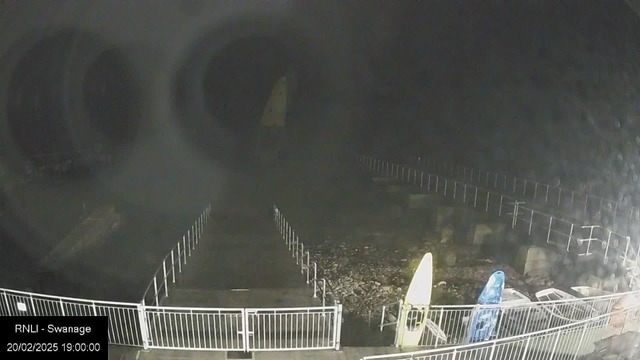 A dark scene captured by a webcam at a coastal location. The foreground features a white fence marking a path leading towards a rocky area. On the right side, two kayaks are visible, one yellow and one blue, near a collection of white containers. The surroundings are shrouded in darkness, with no visible light sources, indicating a nighttime setting.