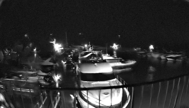 A nighttime scene of a busy marina with multiple boats and yachts docked. The image is predominantly dark with sporadic bright lights reflecting off the water. Several boats are visible in the foreground, with one large white boat taking center stage. The atmosphere appears lively, with the faint outline of more vessels in the background.