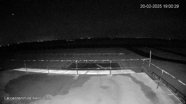 A night-time webcam view showing a flat rooftop with a railing along the edge. There is an open area in the center with a dark surface underneath. In the background, faint lights from distant buildings or vehicles can be seen. The sky is mostly dark with a small bright object, likely a star, visible in the upper left corner. The timestamp in the corner indicates the date and time as 20th February 2025, 19:00:29.