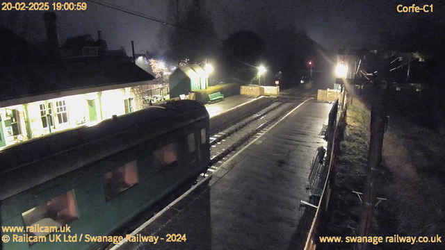 A dark scene of a railway station at night. A green train is partially visible on the left side, with the train tracks extending to the right. The platform is illuminated by a few light sources, revealing a ticket booth and benches. In the background, a small green building with a sloped roof can be seen. The area is surrounded by trees and a wooden fence. The sky is mostly dark, indicating it is evening.