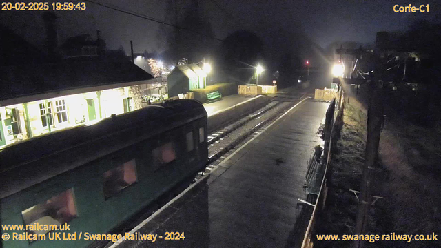 A dimly lit scene of a railway station at night. On the left, a green train is partially visible on the tracks. In the background, the station building has large windows that are illuminated, revealing some interior details. To the right, there is a platform with benches and a small green shelter. Several lights illuminate the area, casting a soft glow. The tracks extend into the distance, leading to a signal light in the background, indicating the station's operational status. The overall atmosphere is quiet and serene.