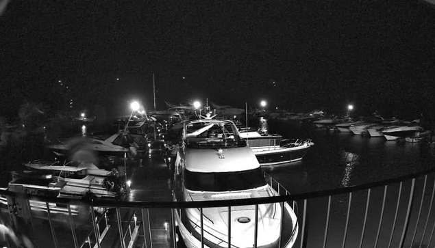 A nighttime scene at a marina with several boats docked in the water. The image is in black and white, displaying bright lights reflecting off the water and illuminating parts of the boats. Some boats are larger yachts, and others are smaller vessels, all docked closely together along a wooden walkway. The atmosphere appears calm and quiet, with the darkness of night surrounding the area.