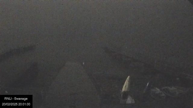 A dark and blurry image taken from a webcam, showing a vague outline of a pier or dock extending into the water, surrounded by fog or low visibility conditions. The setting appears dim, with indistinct shapes suggesting boats or watercraft nearby, and there is a timestamp and identifier at the bottom left corner indicating the date and location.